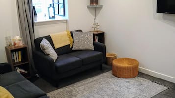Un salon avec un canapé foncé, un petit pouf rond, un tapis à motifs, une bibliothèque et un téléviseur fixé au mur.