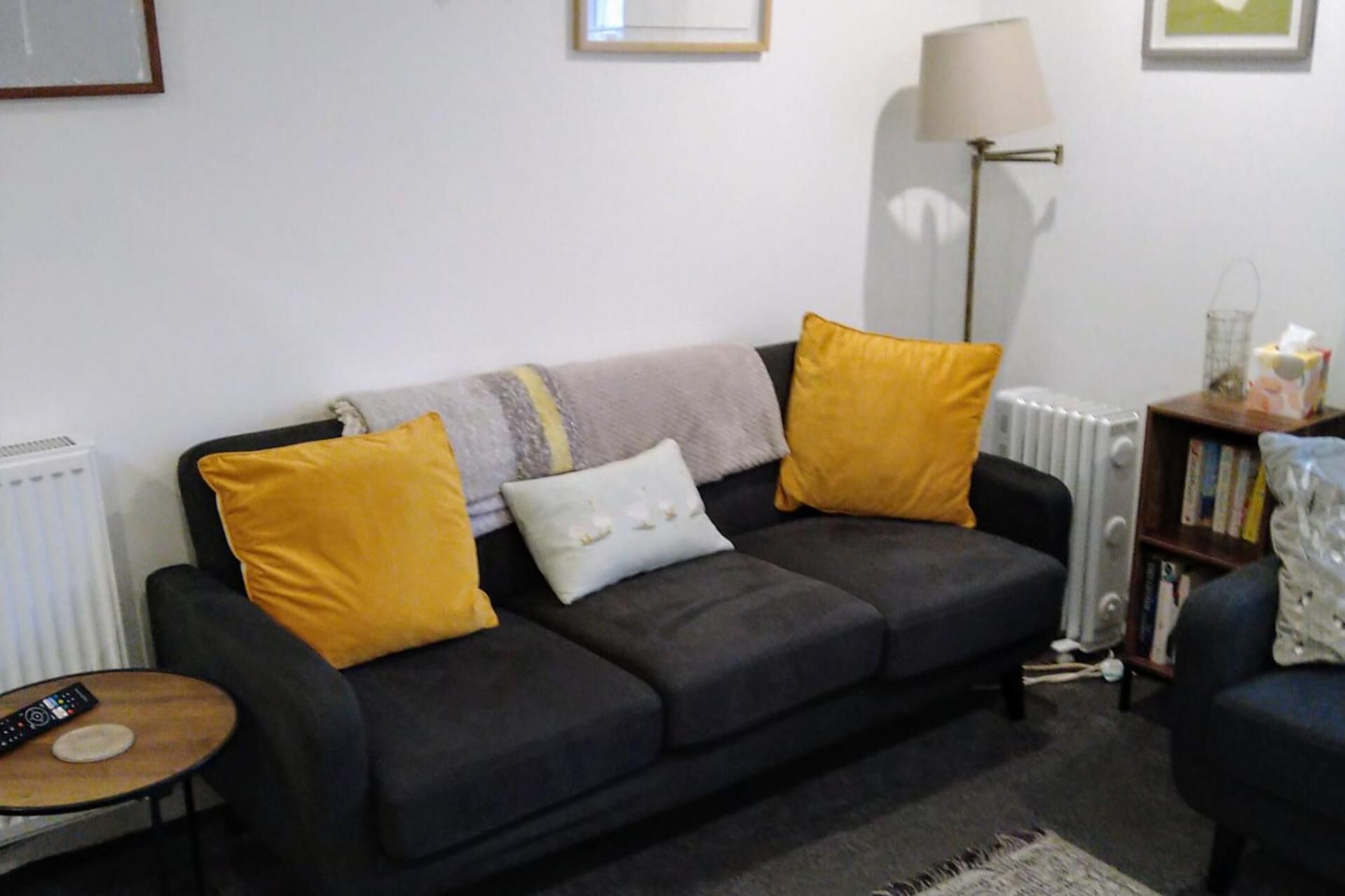 Ein Wohnzimmer mit einem dunklen Sofa, zwei gelben Kissen, einem weißen Kopfkissen, einer Stehlampe, einer Heizung, einem kleinen runden Tisch mit einer Fernbedienung und einem Bücherregal.