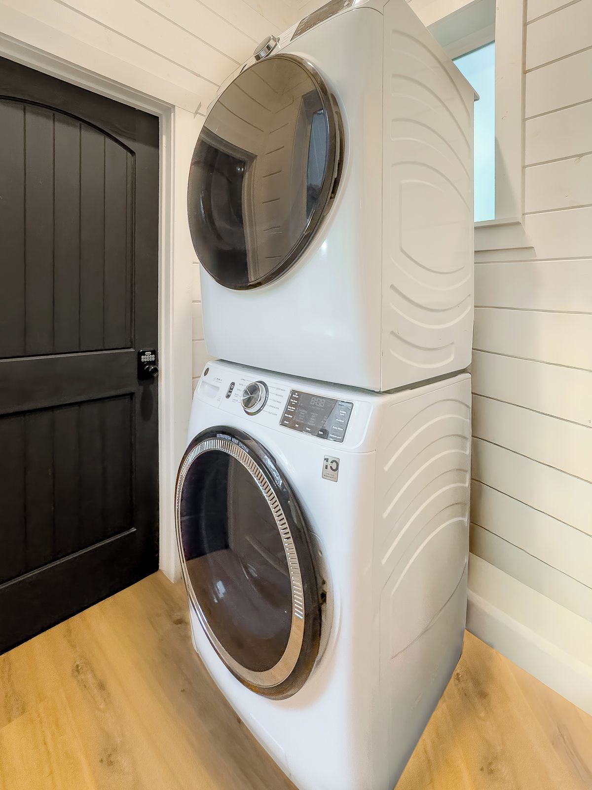 Un appareil combiné lave-linge et sèche-linge à chargement frontal, de couleur blanche, doté d’un panneau de commande et d’un écran numérique, posé sur un plancher en bois, contre un mur blanc.