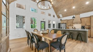 A spacious dining area with a wooden table and chairs, a chandelier, and a view of the outdoors through large windows. - Luxury Treehouse! Elevated, Magical & Unique! (Broken Bow)