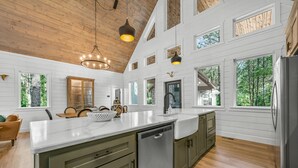 A spacious kitchen with a large island, wooden ceiling, and multiple windows offering views of the outdoors. - Luxury Treehouse! Elevated, Magical & Unique! (Broken Bow)