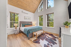 A bedroom with a wooden ceiling, a bed with a blue quilt, a cowhide rug, and a window with a view of trees.