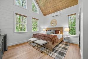 Un dormitorio con techo de madera, ventanas amplias, una cama con almohadas, una mesita de noche con lámpara y un banco.