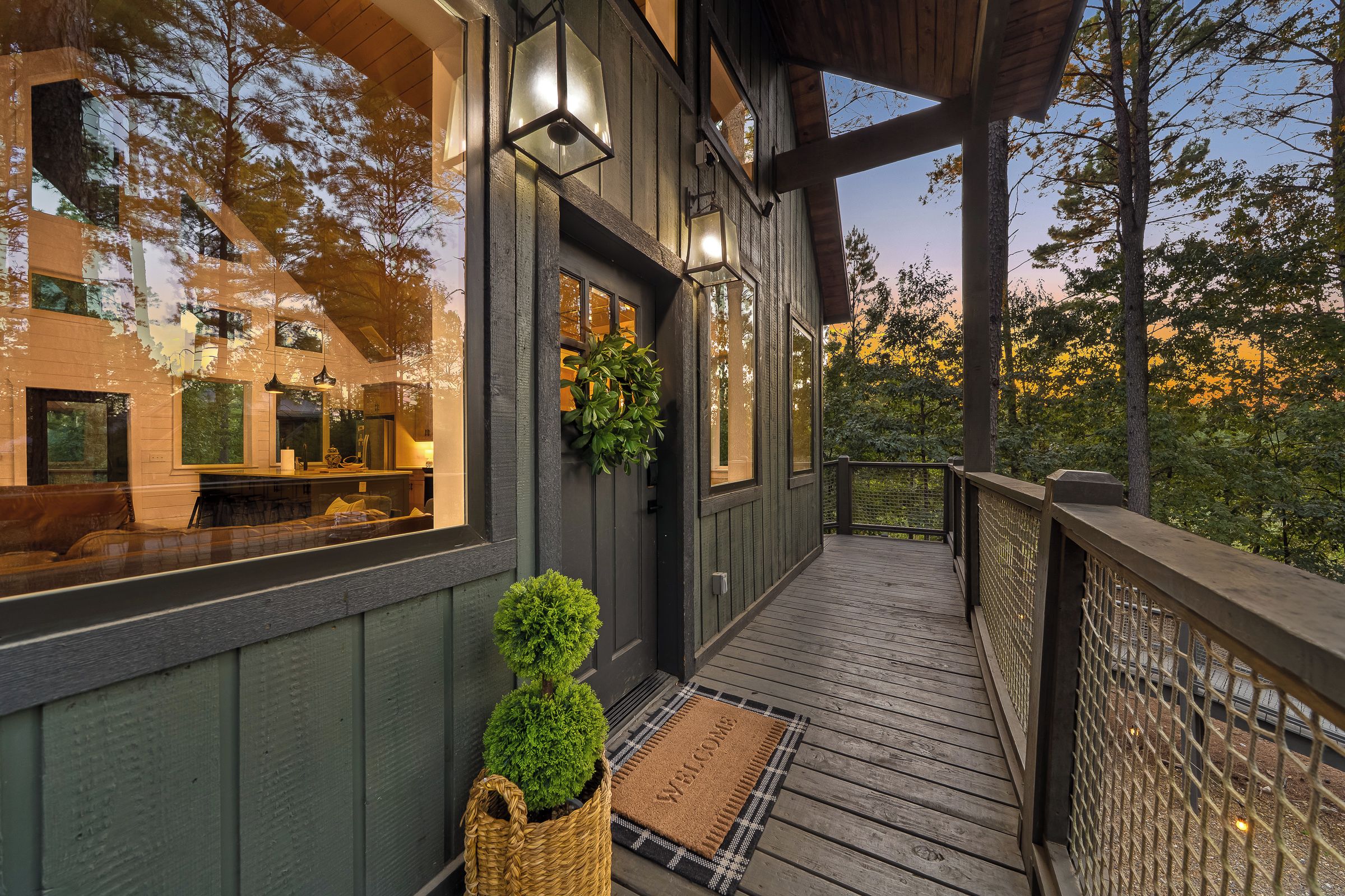A wooden deck with a welcome mat, a potted plant, and a view of the surrounding trees.