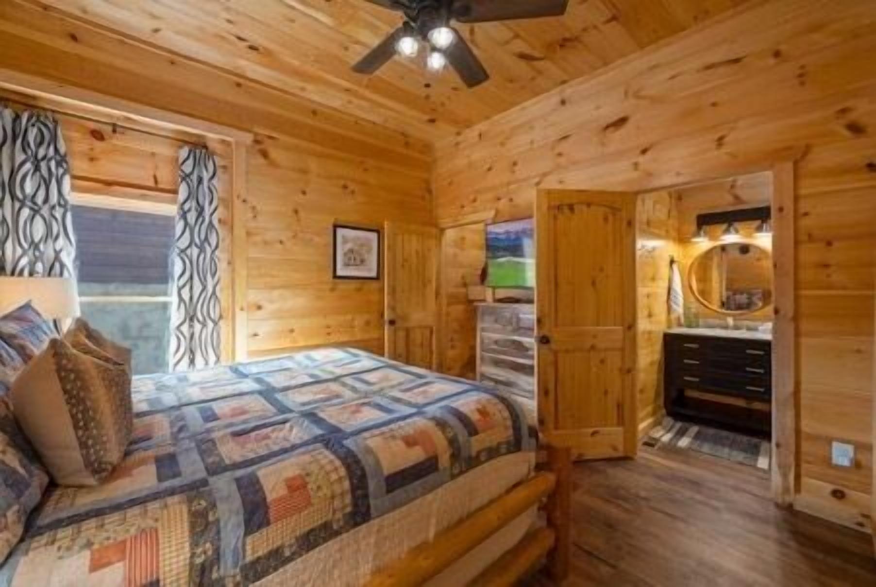Quarto de cabana de madeira com cama, ventilador de teto e banheiro com espelho e pia.