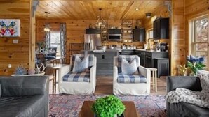 A cozy log cabin interior with a living room, two armchairs, a sofa, a coffee table, and a kitchen area visible through an open doorway. - 🍃Serendipity Lodge🌲Mountain Views, Theater, Game Room & Pet-Friendly (Sevierville)