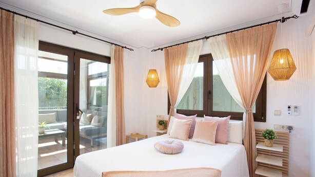 Une chambre à coucher avec un grand lit, des rideaux, un ventilateur de plafond, des tables de chevet et une vue sur l’extérieur, visible à travers une porte vitrée.