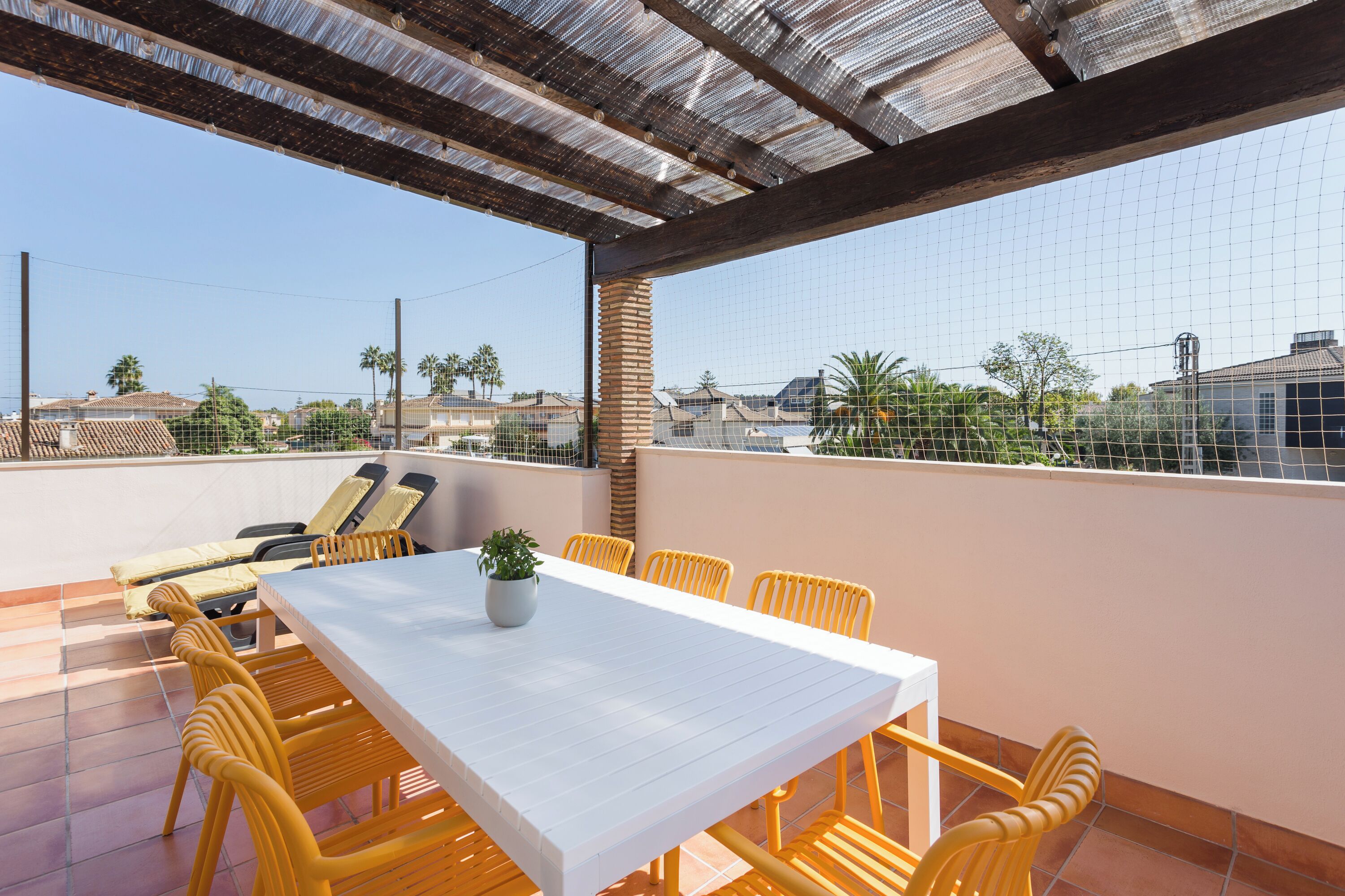 Terraço no telhado com uma mesa branca e cadeiras amarelas, cercado por uma grade de metal e vista para as casas vizinhas.