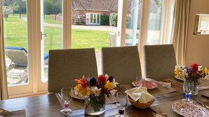 A dining table set for four with chairs, a view of a garden and house through large windows, and a vase with flowers on the table. - The Barn (Hunstanton)