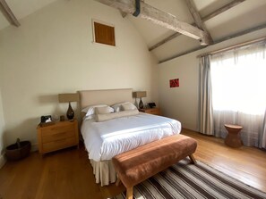 Quarto com cama grande, cabeceira de madeira, mesas de cabeceira com luminárias, um banquinho, uma pequena janela com cortinas e piso de madeira.