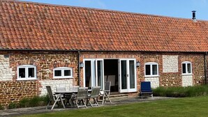 A stone building with a tiled roof, a patio with a table and chairs, and a blue chair. - The Barn (Hunstanton)