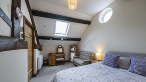 Un dormitorio con techo inclinado, tragaluces, cama con ropa de cama floral, cómoda, silla y espejo.