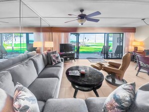 Un salon moderne avec un canapé gris, une table basse ronde et un téléviseur. On y trouve un ventilateur de plafond, une vue sur un paysage verdoyant grâce à de grandes fenêtres, ainsi qu’un coin repas avec des chaises et une table.