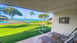 A patio with a view of a well-maintained lawn, a swimming pool, and a building with a framed picture on the wall. - Ma'alaea Surf #G-2 – Relaxing Ground-Floor Getaway (Kihei, Maui)