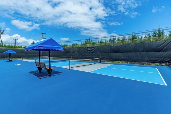Un court de tennis extérieur doté d’une surface bleue, d’un filet et de deux parasols bleus offrant de l’ombre.