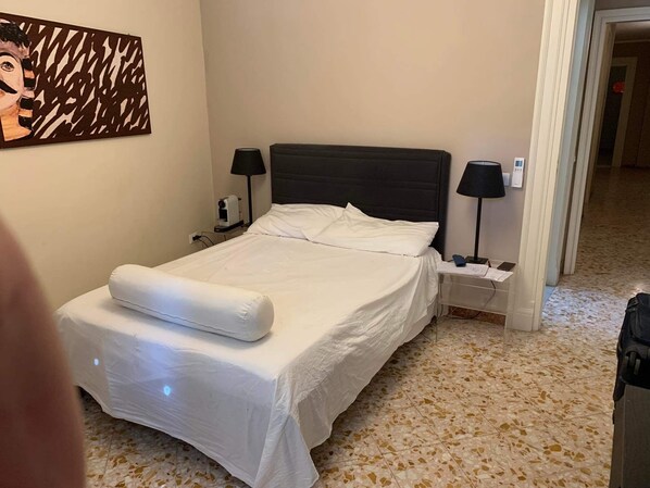 A hotel room with a bed, two bedside tables, a lamp, a painting on the wall, and a tiled floor.