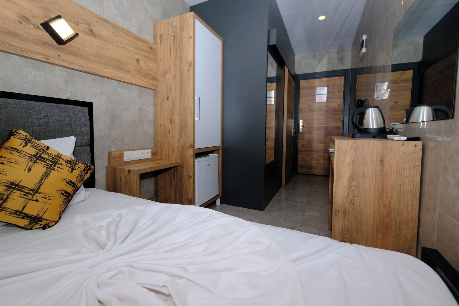 Ein modernes Hotelzimmer mit einem hölzernen Kopfteil, einem Bett mit weißen Leinen, einem kleinen Schreibtisch und einer Küchenzeile mit Wasserkocher und Kaffeemaschine.