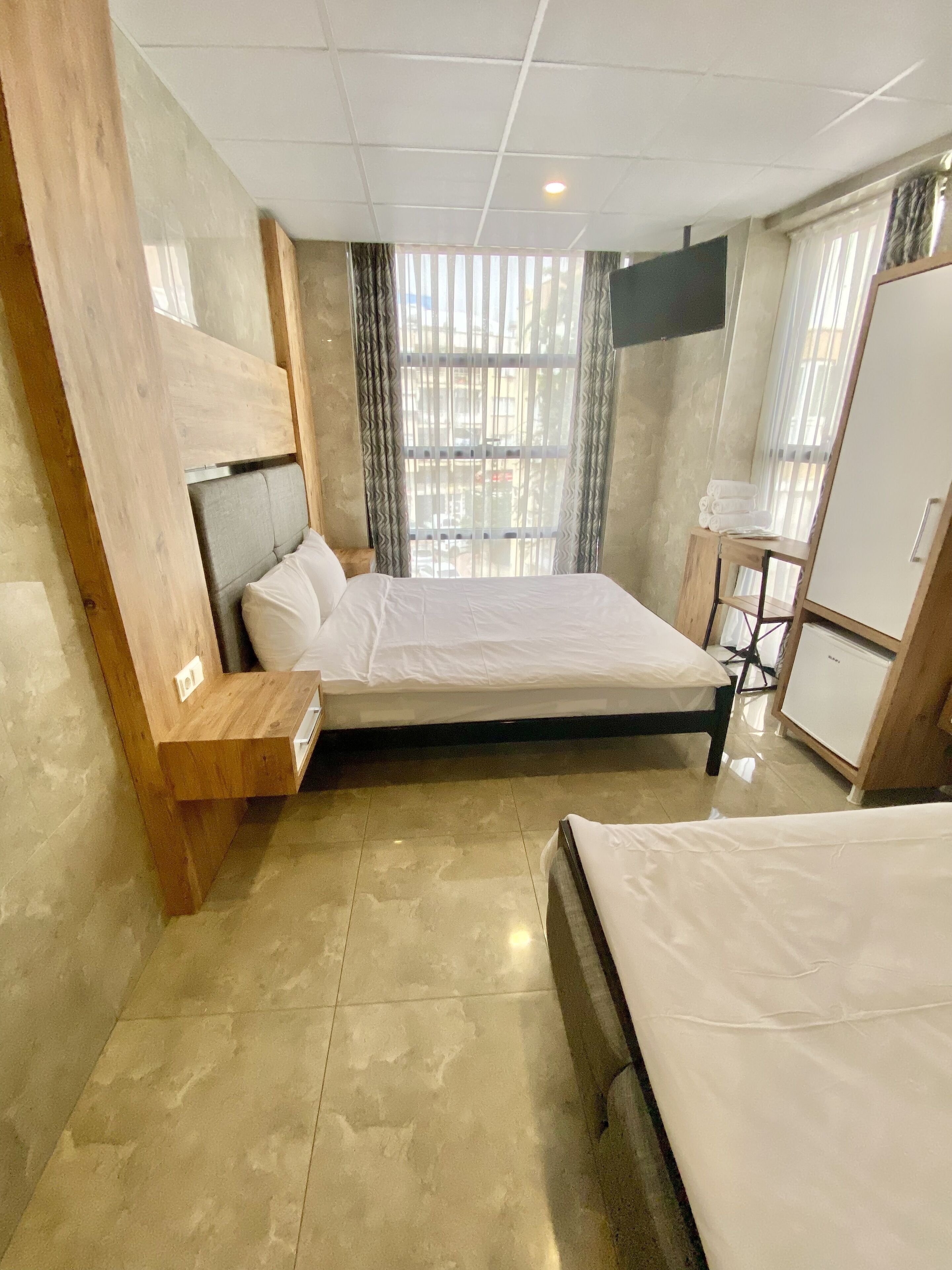 Una habitación de hotel moderna con una cama grande, un cabecero de madera, una mesita de noche, un armario y una ventana con cortinas.