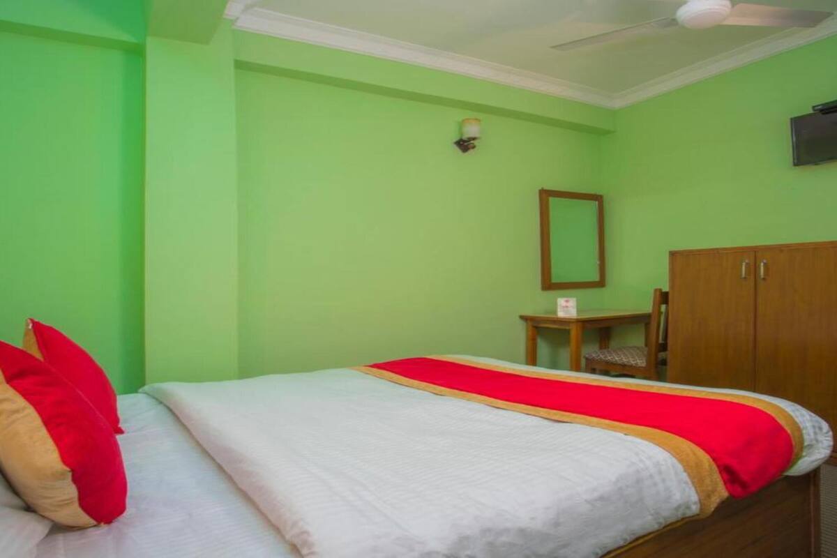 A room with a bed, a wooden wardrobe, a small table with a chair, a wall-mounted TV, and a ceiling fan.