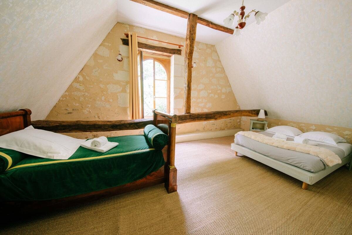 A room with two beds, one with a green bedspread and the other with a white bedspread. There is a wooden beam ceiling and a window with a curtain.