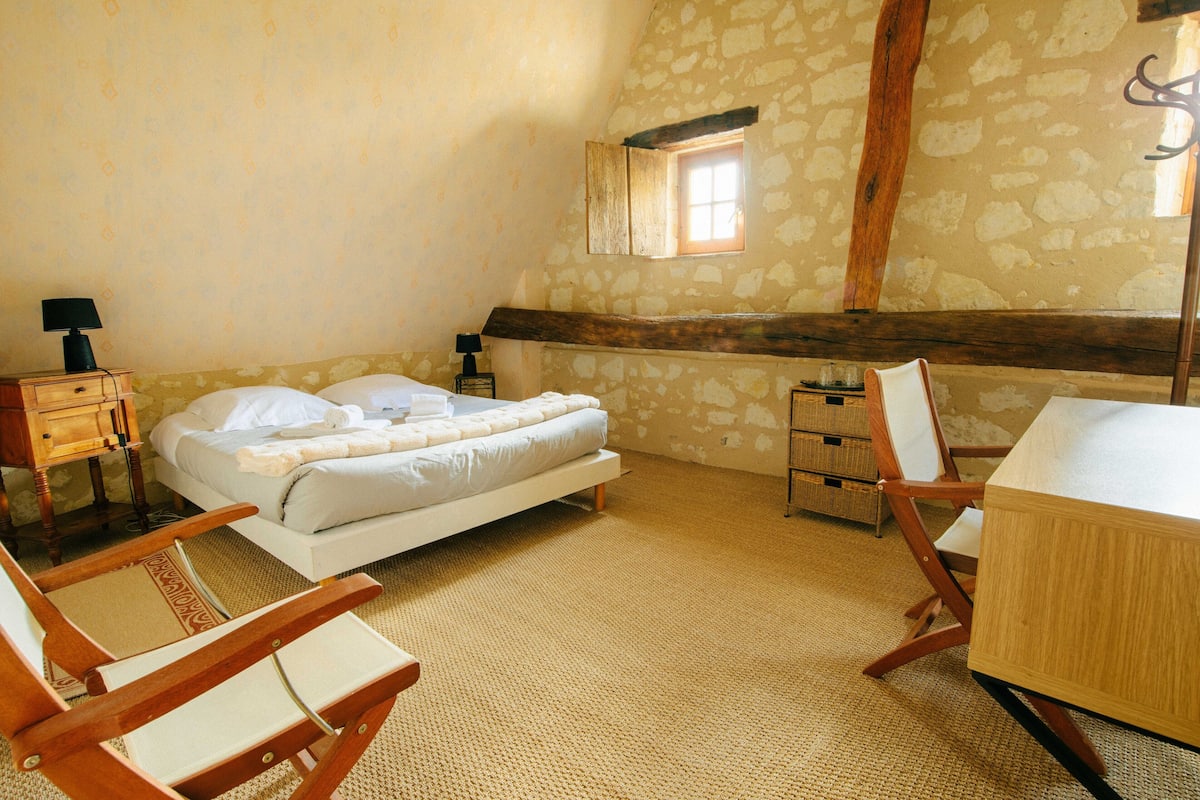 A bedroom with a bed, wooden chairs, a desk, and a stone wall.