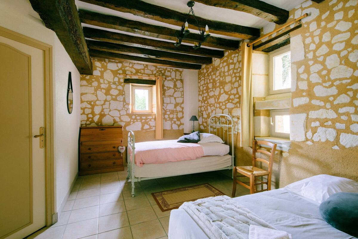 A bedroom with a stone wall, a wooden bed, a wooden dresser, a chair, and a window.