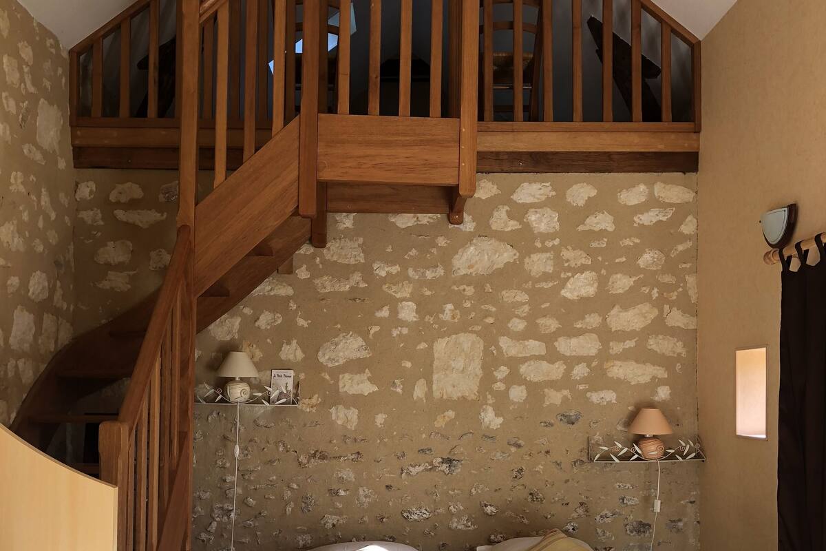A small, cozy room with a wooden staircase, a bed with pillows, a table, and a stone wall.