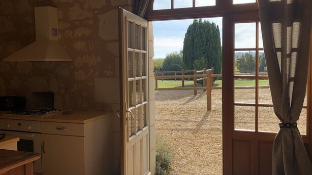 A rustic kitchen with a stone wall, a wooden ceiling, and a large window with a view of a grassy field and trees. - Le Logis de Poëllier (Baugé-en-Anjou)