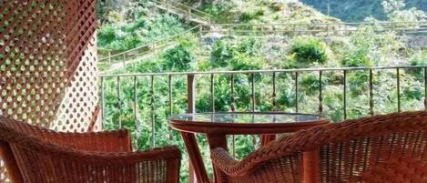 A balcony with wicker chairs and a glass table, overlooking a mountainous landscape.