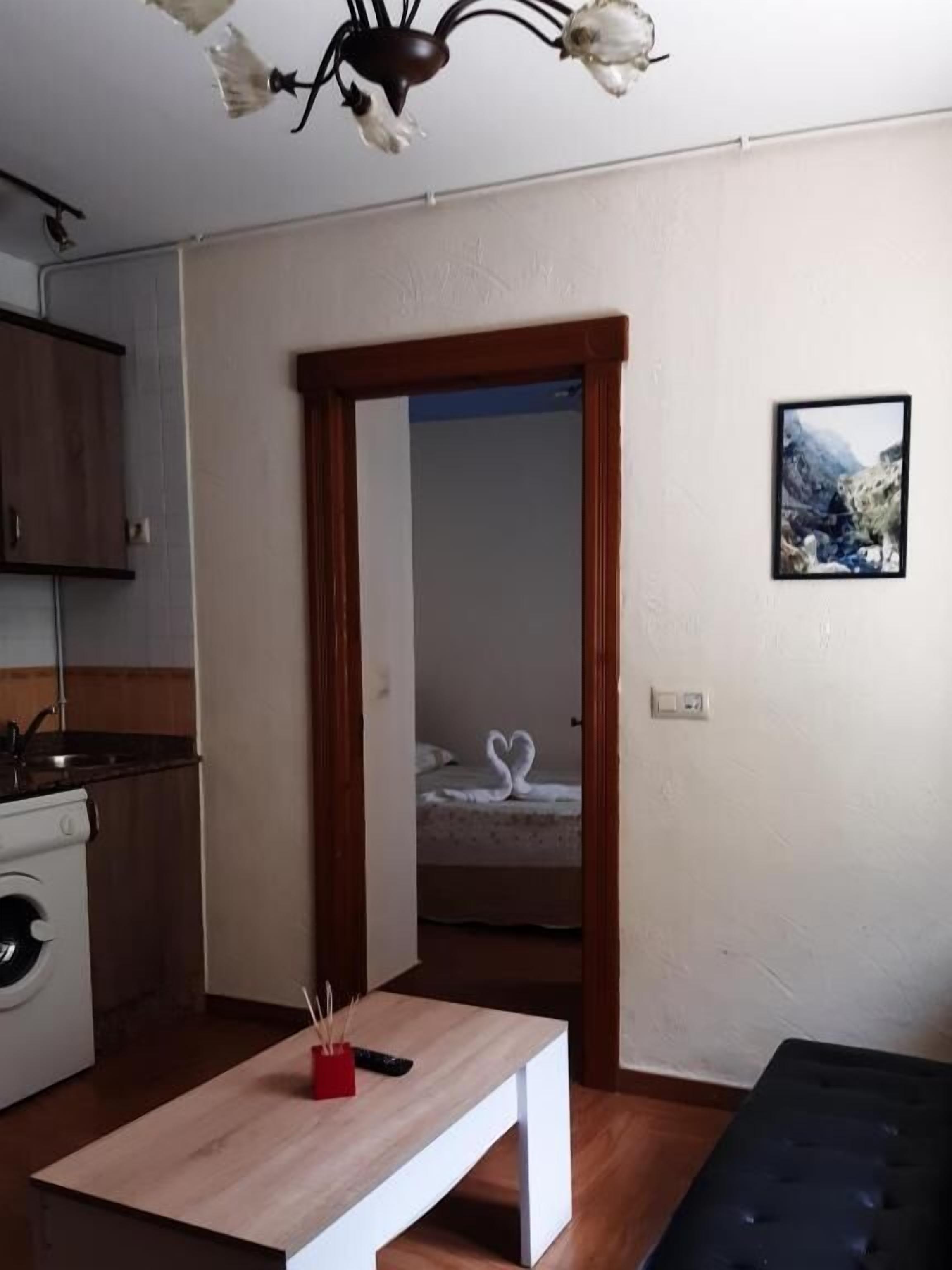 A room with a wooden floor, a white table with a remote control, a bed with a heart-shaped towel, a wooden door, and a picture on the wall.