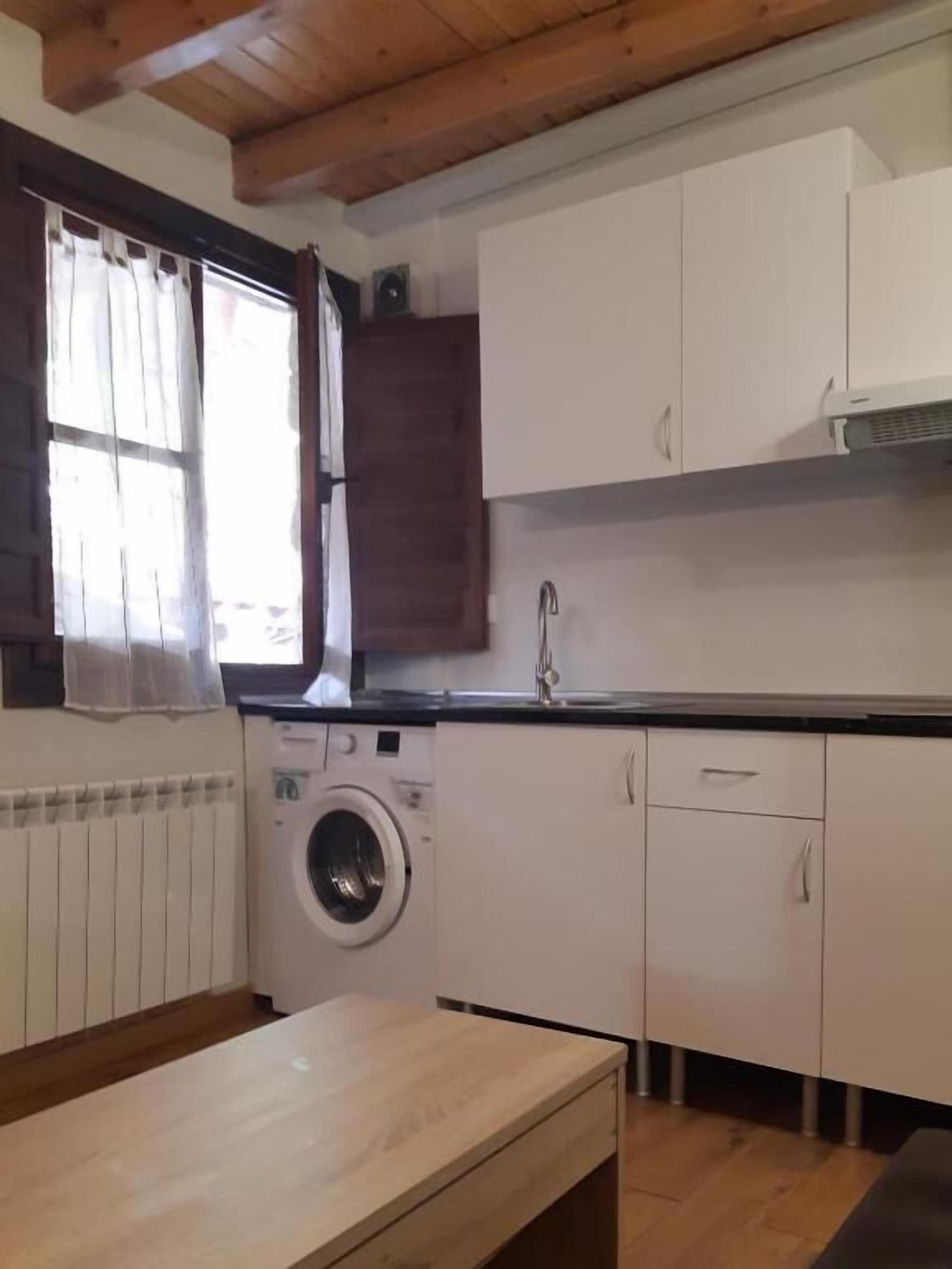 Een kleine keuken met een wasmachine, houten kastjes en een raam met gordijnen.