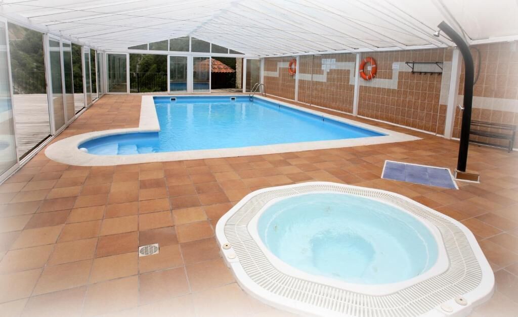An indoor swimming pool with a hot tub, surrounded by glass walls and a tiled floor.