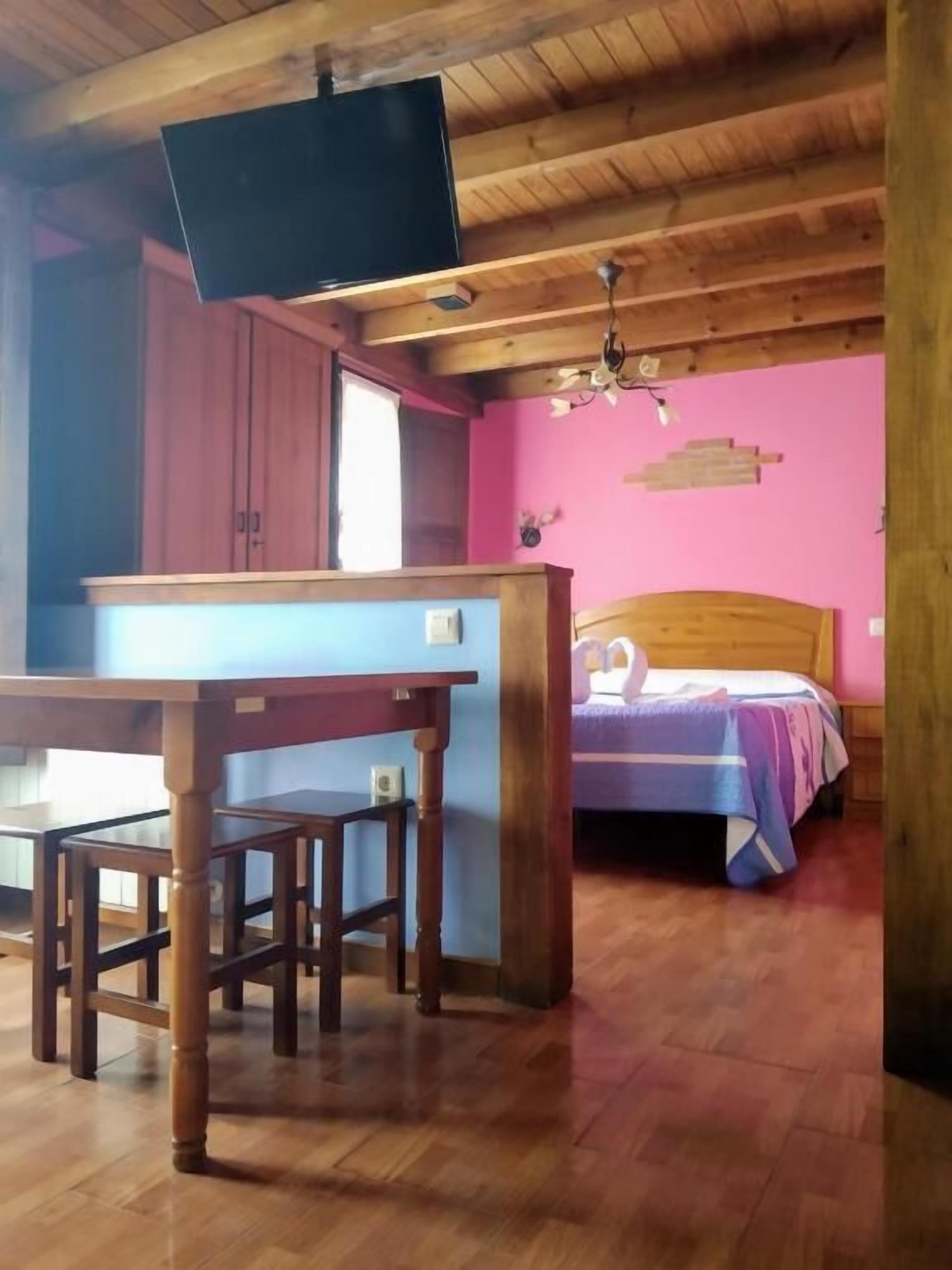 Une pièce avec un plafond en bois, un lit recouvert d’une courtepointe bleue et violette, une table en bois avec des chaises, et un téléviseur fixé au mur.