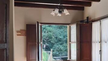 A cozy room with a wooden ceiling, a large window with an open shutter, a sofa, a table, and a chair.