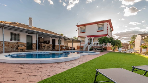 A swimming pool with a stone patio, a grassy lawn, and a house with a red roof and white walls.