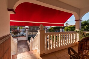 Un balcón con barandilla blanca, toldo rojo y vista a una calle con coches estacionados y áreas verdes.