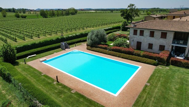 A large rectangular swimming pool surrounded by well-manicured lawns and hedges, with a vineyard in the background and a building with a tiled roof nearby. - Garda & Golf Apartment (Pozzolengo)