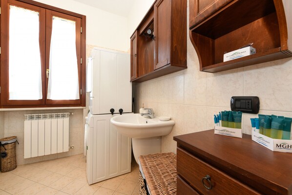 Une cuisine avec des meubles en bois, un évier blanc et un réfrigérateur. On trouve des articles de toilette sur le comptoir et un panier sur le sol.