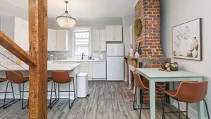 A modern kitchen with white cabinets, a brick wall, and a dining area with a light blue table and brown chairs. - Historic NoDa Mill House • Steps to Shops & Dining (Charlotte)