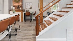 A modern kitchen with a brick accent wall, a dining area with wooden chairs, and a staircase with wooden railings. - Historic NoDa Mill House • Steps to Shops & Dining (Charlotte)