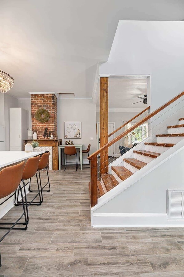 A modern kitchen with a brick accent wall, a dining area with wooden chairs, and a staircase with wooden railings. - Historic NoDa Mill House • Steps to Shops & Dining (Charlotte)