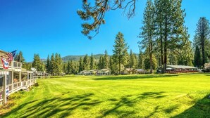 A well-maintained grassy area with several buildings, including a lodge with a deck and an American flag, surrounded by tall pine trees and a clear blue sky. - Caddyshack - 35 Aspen Circle (Plumas Eureka)
