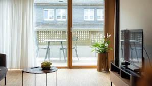 Een moderne woonkamer met een schuifglasdeur naar een balkon, een televisie, een klein rond tafeltje met een fruitschaal, en een potplant.