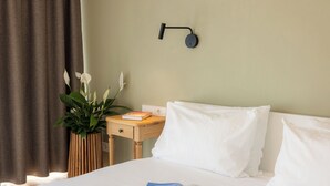 Een net opgemaakt bed met witte lakens, een nachtkastje met een boek en een plant in pot, en een wandlamp.