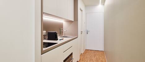 A narrow hallway with a kitchen area on the left, featuring white cabinets, a countertop, and a sink.