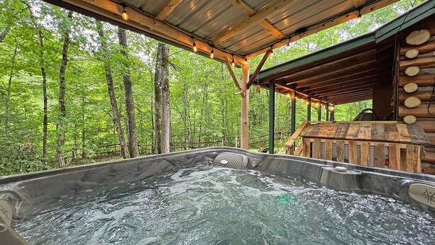 Een hottub met een houten terras en een metalen dak, omgeven door een bos.