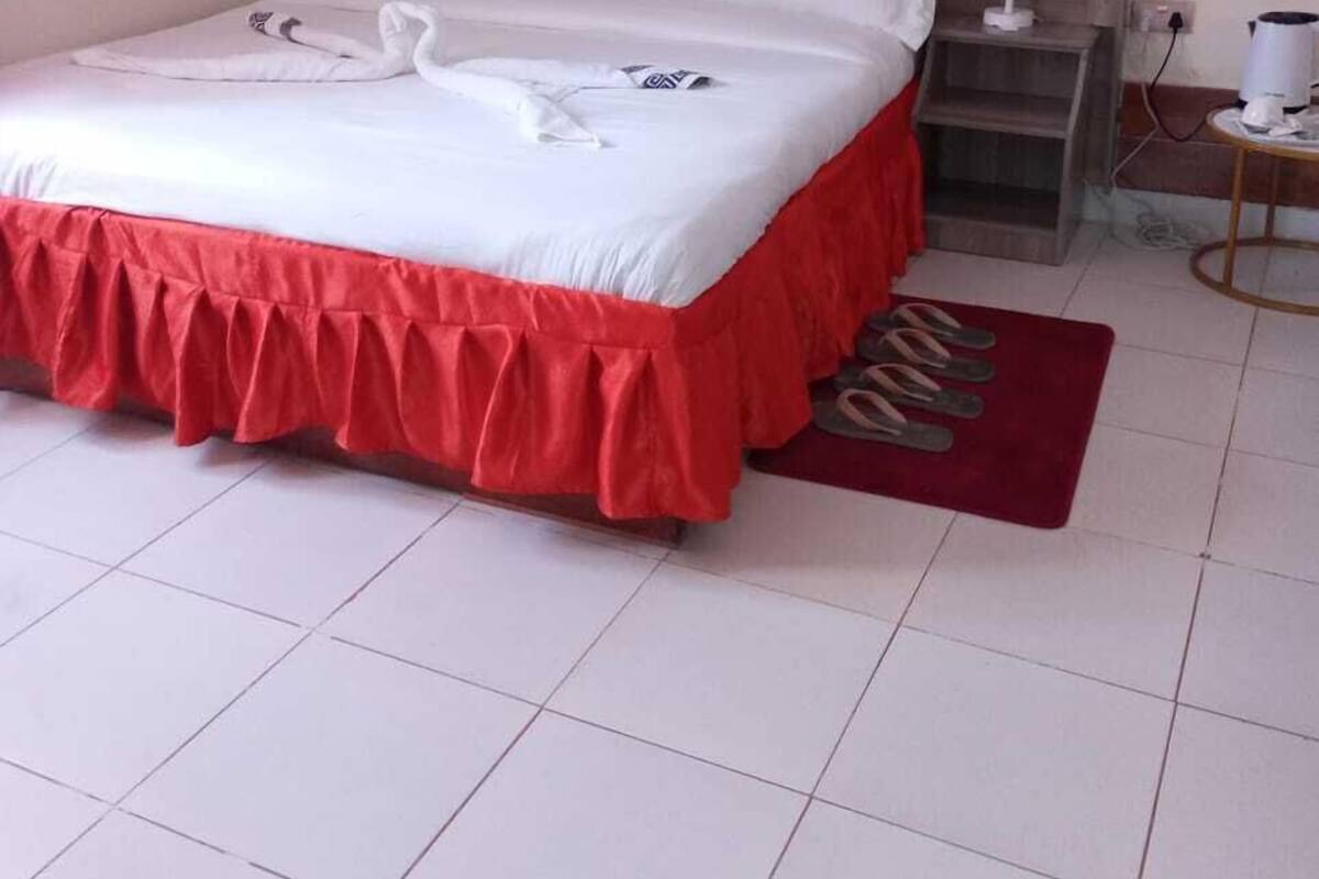 Uma cama bem arrumada com saia vermelha, uma mesinha de cabeceira com abajur, e um tapete vermelho em piso de azulejo.