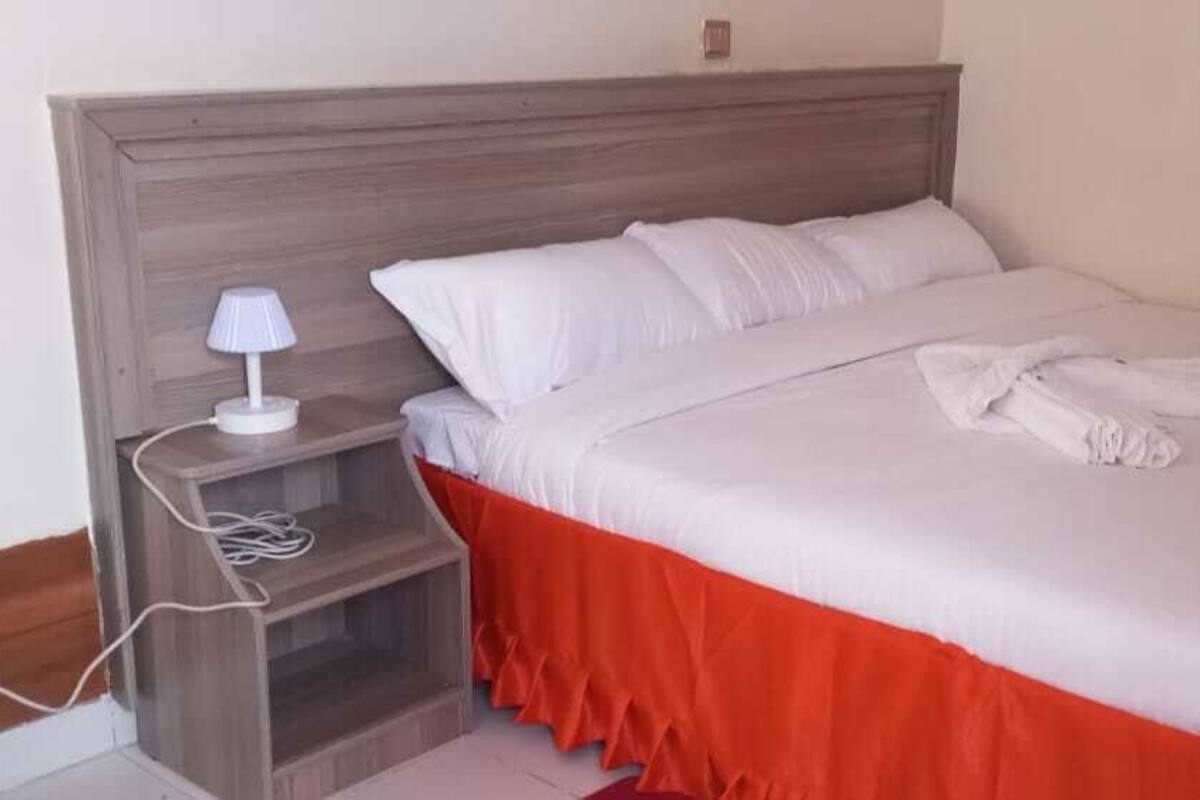 Cama bem arrumada com roupa de cama branca e saia vermelha, mesa de cabeceira com abajur e uma tomada.