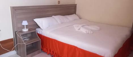 A neatly made bed with white bedding and a red skirt, a bedside table with a lamp, and a power outlet.