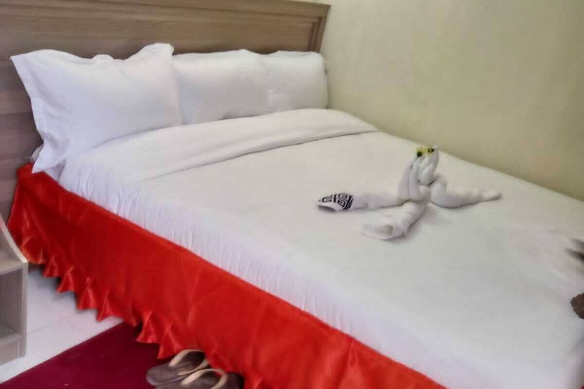 Uma cama bem arrumada com roupa de cama branca e vermelha, um par de chinelos sobre um tapete vermelho e um brinquedo de pelúcia na cama.